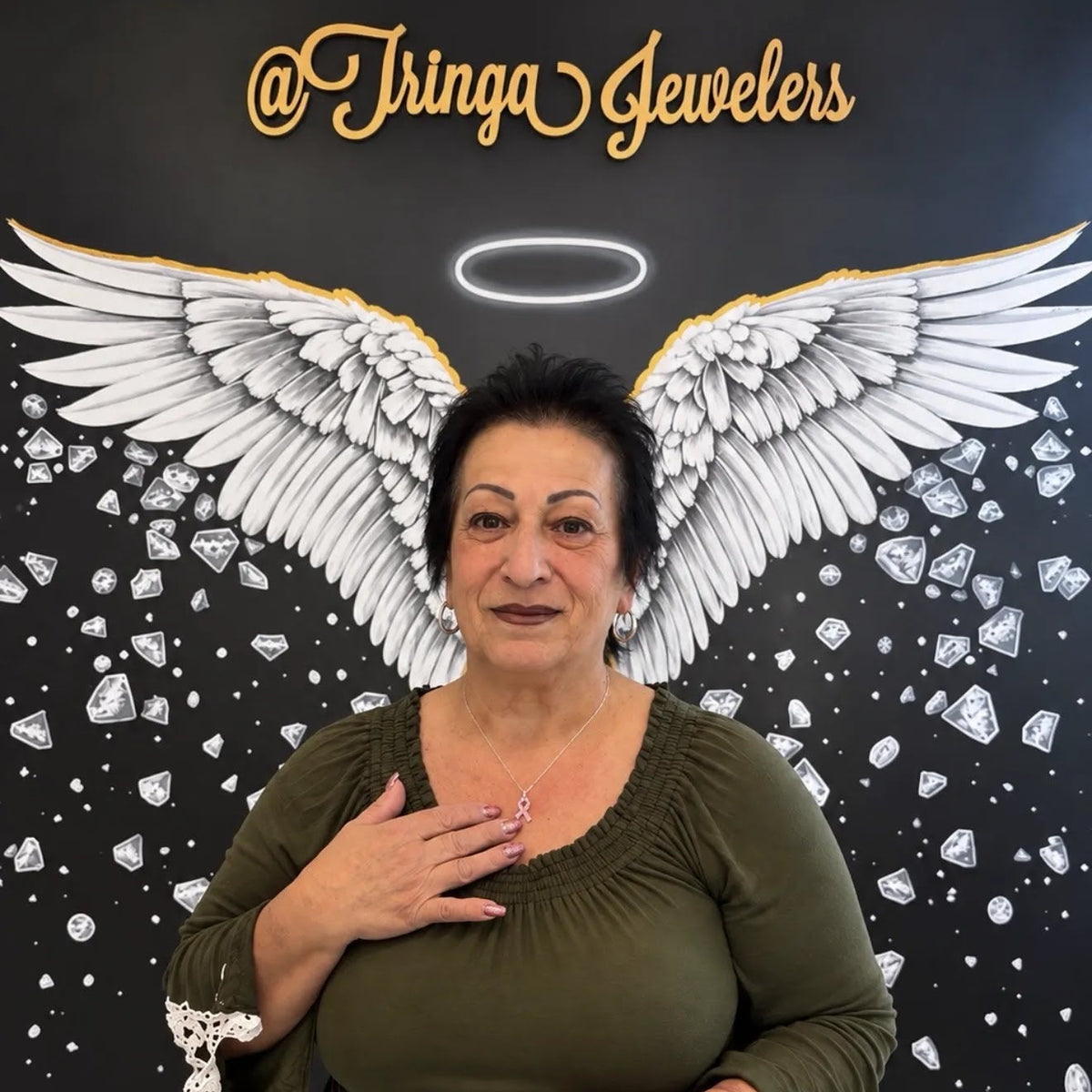 A woman wearing an olive green long-sleeve top with white lace cuffs stands in front of a black wall decorated with large white angel wings, a halo, and scattered diamond graphics. The text '@Tringa Jewelers' is visible at the top.