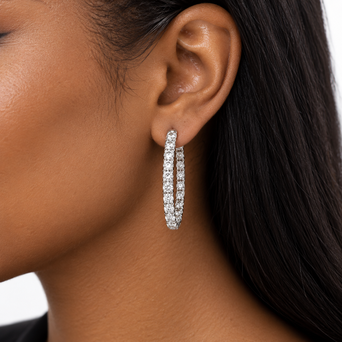 A close-up of a model's ear wearing a silver hoop earring set with small clear stones, shown against dark hair and a light background.