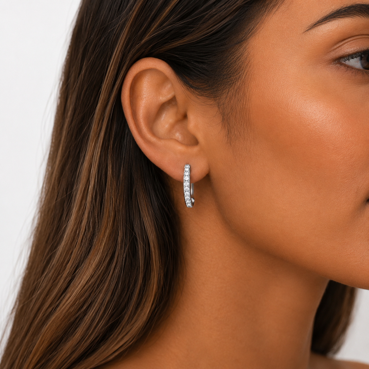 A close-up of a woman's ear wearing a small silver hoop earring set with clear stones, against a clean light gray background.
