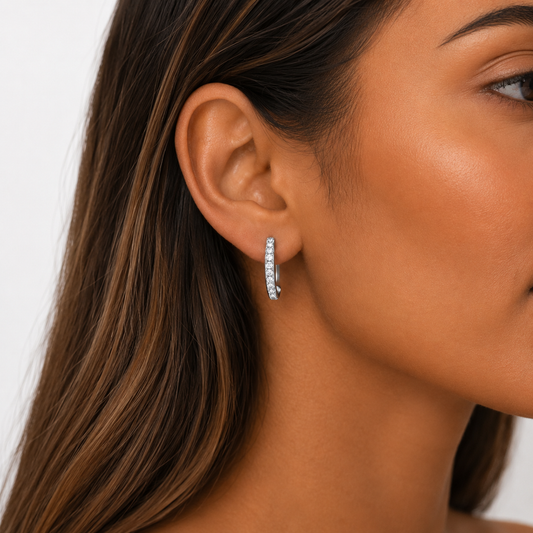 A close-up of a woman's ear wearing a small silver hoop earring set with clear stones, against a clean light gray background.